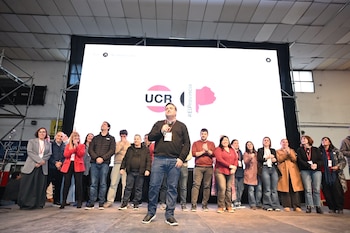 Un hombre habla con un micrófono en un escenario. Hay personas de pie a su alrededor. Detrás se ve una pantalla grande con el logo UCR y un mapa rosado