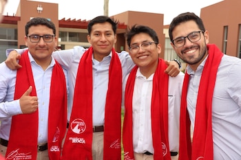 Cuatro hombres jóvenes con lentes y camisas sonríen abrazados en el exterior de un edificio mientras llevan bufandas rojas