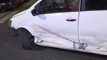 Un coche blanco muestra el guardabarros delantero y las puertas del lado izquierdo severamente abollados y rayados tras un impacto, con el pavimento y otros vehículos oscuros al fondo