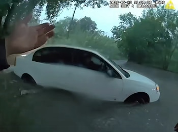 Un coche blanco está sumergido hasta la mitad en aguas de inundación en una carretera, mientras una mano se extiende hacia él. Hay árboles y cielo nublado de fondo