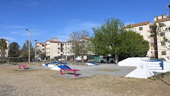 Una calle rodeada de edificios residenciales, un espacio de tierra con césped seco, rampas de skate, bancos, árboles y vehículos estacionados
