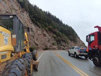 Carretera mojada con doble línea amarilla, maquinaria pesada amarilla, camión comercial rojo, camioneta pickup blanca, montaña con árboles y rocas