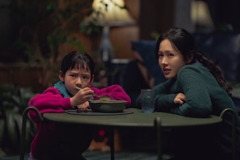 Una mujer asiática y una niña sentadas a una mesa oscura. La niña, en suéter fucsia, come de un cuenco. La mujer, con suéter verde oscuro, mira