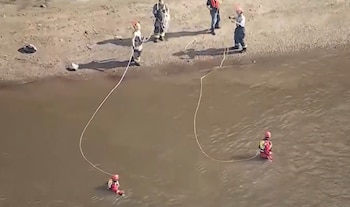 Cuatro rescatistas en la orilla sostienen cuerdas sujetas a dos miembros del equipo desplegados dentro del agua de un río
