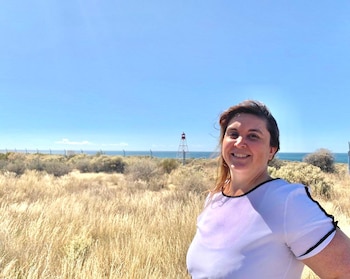 Mujer sonriente con camiseta blanca, vegetación seca, el mar, un cielo azul, una torre rojiblanca y una valla al fondo