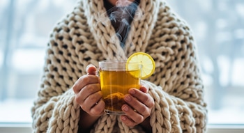 Manos sosteniendo una taza de té de jengibre humeante con una rodaja de limón, una persona con una manta de punto grueso, y una ventana invernal borrosa al fondo.