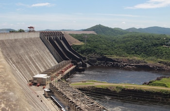 Represa de hormigón con estructura de compuertas y plataforma, agua oscura fluyendo abajo, colinas verdes y cielo azul con nubes blancas