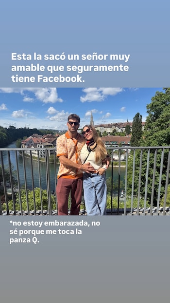 Mujer y hombre sonrientes abrazados sobre un puente metálico. Detrás, un río, edificios antiguos con tejados y abundante vegetación, bajo cielo azul claro