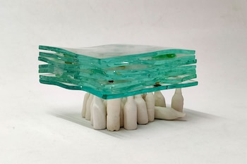 Escultura abstracta compuesta por varias capas de vidrio verde translúcido apiladas sobre múltiples piezas de cerámica blanca con forma de botella