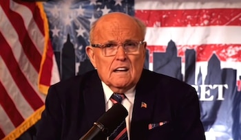 Rudy Giuliani habla frente a un micrófono con gafas y traje oscuro, ante un fondo con la bandera de Estados Unidos