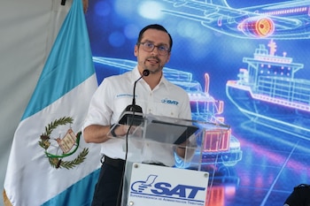 Hombre con gafas en podio transparente, bandera de Guatemala, logotipo SAT, pantalla digital azul con esquemas de avión, barco y camión