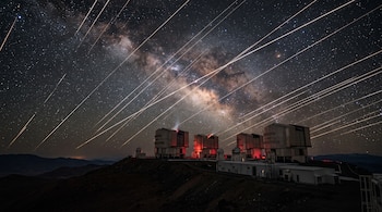 Edificios de un observatorio astronómico en una montaña bajo un cielo estrellado cruzado por trazos luminosos.