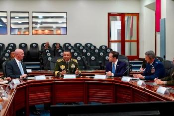 Mesa ovalada de madera, cinco hombres sentados (tres en uniforme militar, dos en traje). Un hombre militar gesticula. Sillas vacías detrás. Nameplates