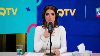 Una mujer con auriculares y micrófono en una mesa con un vaso de agua, documentos y una caja de pañuelos, con pantallas de televisión al fondo.