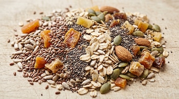 Un montón de semillas de chía, lino, girasol y calabaza mezcladas con copos de avena, almendras y trozos de fruta deshidratada sobre madera.