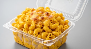 Primer plano de una porción de macarrones con queso amarillo intenso dentro de un recipiente plástico transparente abierto, con la superficie gratinada y puntos de pimienta negra.