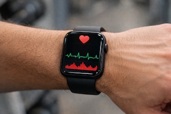 Primer plano del brazo de una persona con un reloj inteligente negro mostrando un gráfico de línea verde y barras rojas para análisis cardíaco y un corazón rojo en la pantalla.