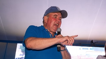 Un hombre con gorra azul y camisa polo azul habla por un micrófono mientras señala con la mano derecha.