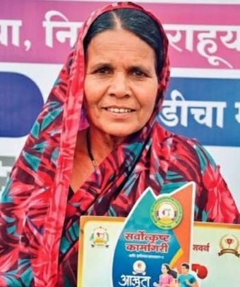 Una mujer con túnica estampada roja sostiene un certificado impreso con letras en escritura devanagari y un trofeo