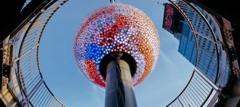 Vista en contrapicado de la esfera de Times Square, cubierta de luces multicolores. Una barandilla curva la enmarca, con el edificio One Times Square detrás