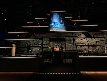Una sala de exhibición oscura presenta un monitor con un tigre digital azul brillante, flanqueado por vitrinas con objetos y paredes con gráficos de ruinas antiguas