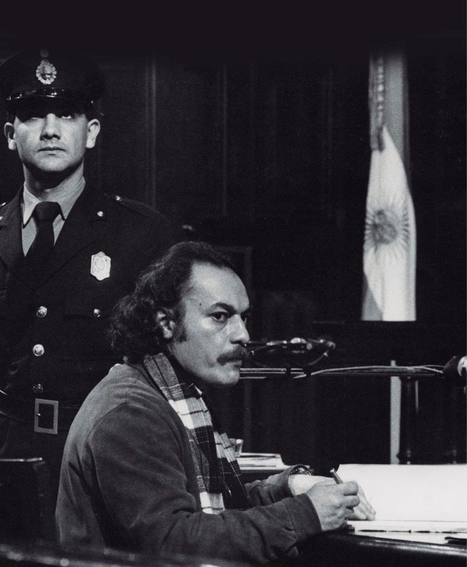 Víctor Basterra, sobreviviente y testigo clave de la dictadura argentina, redacta su declaración en un tribunal bajo la atenta mirada de un oficial, con la bandera argentina visible al fondo. (Jorge Sánchez)