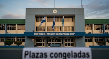 Edificio gris, azul y crema del Ministerio de Hacienda y Crédito Público. Tres banderas de Nicaragua, una banca azul y un cartel con 'Plazas congeladas'.