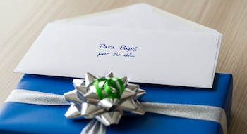 Primer plano de un sobre blanco con "Para Papá por su día" en tinta azul, sobre un regalo azul con cinta plateada y moño decorativo verde, en mesa de madera clara.