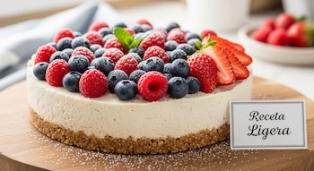 Tarta de queso con base de galleta, crema blanca y frutos rojos (frambuesas, arándanos, fresas) espolvoreados con azúcar glas, junto a una tarjeta 'Receta Ligera'.