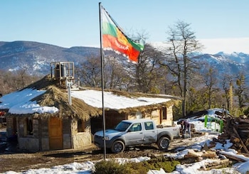 Grupo de personas Mapuche alrededor de una fogata, con un tambor, frente a pistas de esquí nevadas y un remonte en el Cerro Chapelco, bajo un cielo azul. Una bandera Mapuche flamea a la derecha