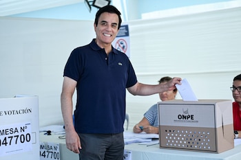 Carlos Espá, sonriente, viste un polo azul y deposita una papeleta en una urna de votación marrón con el logo de ONPE, con otras personas al fondo