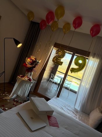 Habitación de hotel decorada para un cumpleaños: globos con el número 29, flores, pétalos de rosa, portátil en la cama y un balcón con vista al lago