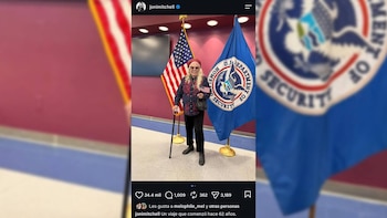 Una mujer sostiene un bastón y una pequeña bandera frente a la bandera de Estados Unidos y el sello del Departamento de Seguridad Nacional