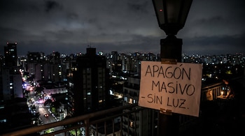 Cartel blanco con "Apagón masivo - Sin luz" en farol, con edificios y luces urbanas de ciudad nocturna. Una persona usa una vela.