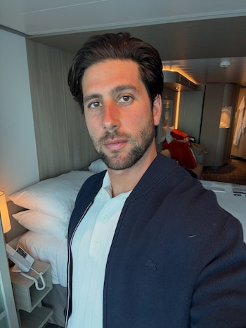 Hombre con barba y pelo oscuro viste polo blanco y chaqueta azul con logo de cocodrilo. Fondo con cama, almohadas blancas, mesa de noche y teléfono