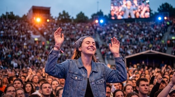 Mujer con chaqueta vaquera y pañuelo levanta los brazos en concierto. Multitud de personas. Pantalla y luces en segundo plano.