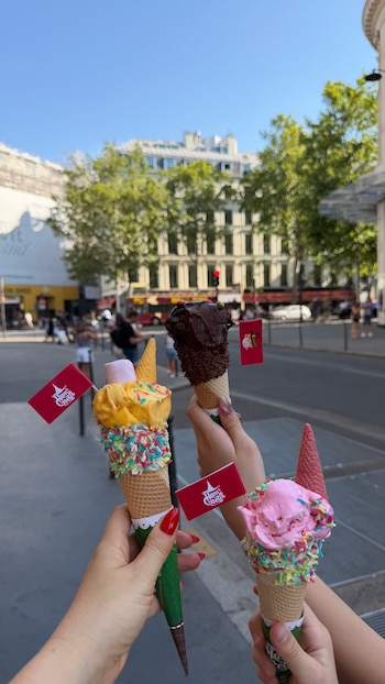 Tres manos sostienen cucuruchos de helado con diferentes sabores, grageas y malvaviscos. Dos tienen banderines rojos con un logo blanco. Fondo de calle y edificios de ciudad