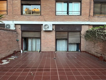 Patio de baldosas rojas con suciedad y algunas rotas, frente a una pared de ladrillo con puertas correderas, persianas, y dos unidades de aire acondicionado