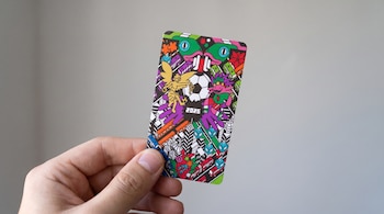 Mano sosteniendo una tarjeta rectangular con diseño de figuras coloridas, un balón de fútbol, el número 2026 y un fondo gris claro.