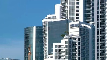 Una vista de varios edificios altos modernos con fachadas blancas y de vidrio contra un cielo azul claro. Un brazo de grúa de construcción es visible a la izquierda