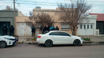 Calle con vehículos, incluyendo un coche policial y un auto civil blanco, y un grupo de policías en uniformes y equipos tácticos frente a varias casas