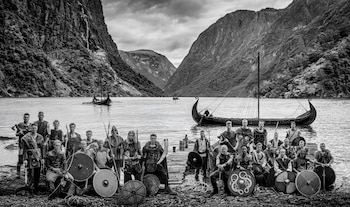 Un grupo de hombres vestidos como vikingos posa junto a un fiordo noruego, con barcos largos y montañas de fondo