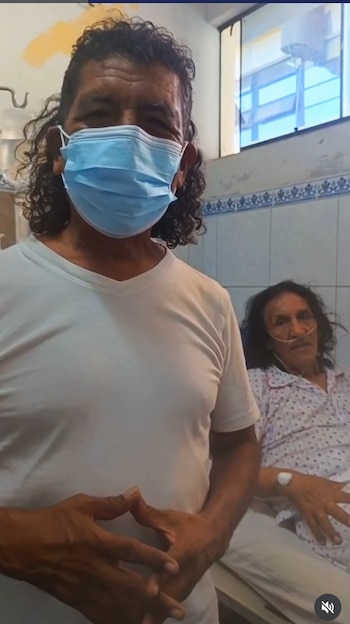 Un hombre con mascarilla azul y camiseta blanca posa de pie junto a un hombre acostado en una cama de hospital