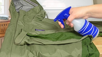 Una mano sostiene un pulverizador azul y rocía una chaqueta verde extendida sobre una mesa de madera.