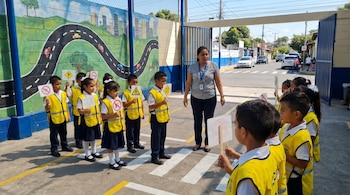 Niños con chalecos amarillos y una adulta practican seguridad vial en un cruce peatonal simulado, con un mural de carretera de fondo. Los niños sostienen señales de tráfico.
