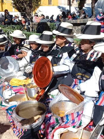 Varias mujeres y un niño sentados al aire libre con sombreros blancos y negros, vestimenta tradicional, y ollas de comida sobre telas coloridas