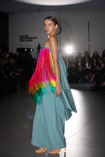 Una modelo viste un traje largo de color azul con franjas de flecos fucsia, amarillo y verde sobre una pasarela de exhibición