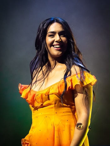 Angela Leiva sonríe vestida con un traje amarillo de volantes y hombros descubiertos sobre el escenario