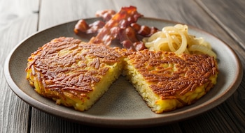 Rösti suizo dorado y cortado por la mitad en un plato de cerámica con tocino crujiente y cebolla salteada sobre una mesa de madera.