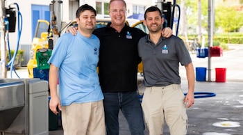 Tres hombres posan sonrientes frente a un SUV compacto amarillo en un lavadero de autos. El central viste polo negro, el de la izquierda camiseta azul claro y el de la derecha polo gris